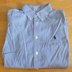 Randig skjorta från Ralph Lauren - Snygg blå- och vitrandig skjorta från Ralph Lauren med klassisk krage och knappar framtill. Skjortan har lång ärm och ikonisk broderad logga på bröstet. Perfekt för dig som gillar stilrena och tidlösa plagg.