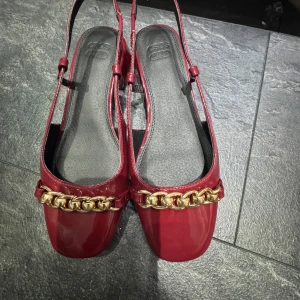 Vinröda ballerinaskor med kedjedetalj - Vinröda ballerinaskor från ASOS Design med lackad finish, fyrkantig tå och chunky guldfärgad kedja framtill. Skorna har öppen häl med justerbara remmar och låg klack. Perfekta för dig som vill sticka ut med en trendig och elegant look.