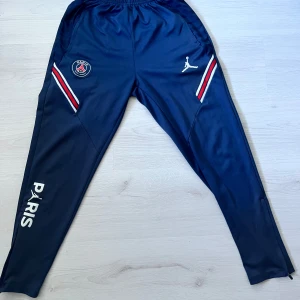 PSG x Jordan träningsbyxor - Marinblå träningsbyxor från PSG x Jordan med vita och röda detaljer vid fickorna, PSG-logga och Jumpman-logga framtill samt PARIS-tryck på benet. Byxorna har elastisk midja och smal passform, perfekta för fotboll eller chill.