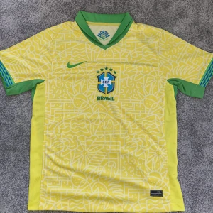 Brasilien fotbollströja Nike gul/grön - Snygg Brasilien fotbollströja från Nike i Dri-FIT-material. Tröjan är gul med gröna detaljer och har ett unikt grafiskt mönster över hela ytan. Klassisk V-ringad krage i grönt, korta ärmar och officiellt CBF-märke på bröstet. Perfekt för dig som älskar fotboll och vill sticka ut.