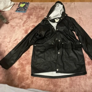 Svart regnjacka från 157 XS - Svart regnjacka från 157 i storlek XS. Jackan har huva med dragsko, stora fickor med lock och tryckknappar framtill. Perfekt för regniga dagar med sin vattenavvisande yta och klassiska look. Enkel att matcha och riktigt praktisk.