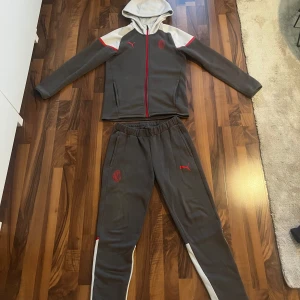  AC Milan träningset  - Säljer ett par grå AC Milan mjukisbyxor från Puma med röda detaljer och vita paneler längst ner på benen. Byxorna har resår i midjan, smal passform och är perfekta för träning eller chill. AC Milan-logga och Puma-logga framtill.