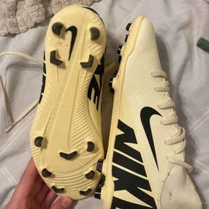 Beige Nike fotbollsskor med svarta detaljer helt ny - Snygga beige fotbollsskor från Nike med stora svarta loggor på sidorna och svarta dobbar. Skorna har snörning och är tillverkade i syntetmaterial för lätt känsla på planen. Perfekta för dig som vill sticka ut med stilren design och bra grepp.