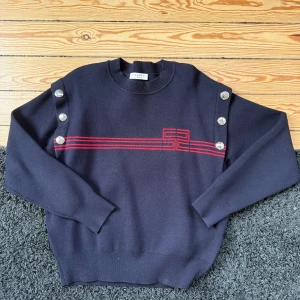 Marinblå tröja med röda ränder från Sandro - Svart långärmad tröja från Sandro som är oversize med röda linjer och grafiskt mönster framtill. Snygga silverfärgade knappar längs axlarna ger en unik touch. Materialet är en mjuk mix av viskos, polyester och polyamid. Perfekt för dig som gillar stilrena detaljer och cool design.