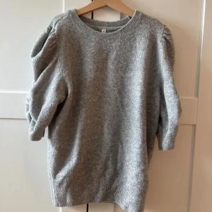 Grå stickad tröja med puffärm - Mysig grå stickad tröja i storlek S med puffade trekvartsärmar och rund hals. Tröjan har ribbade muddar vid ärmslut och nederkant, vilket ger en snygg och chill look. Perfekt för dig som gillar enkel stil med en twist.