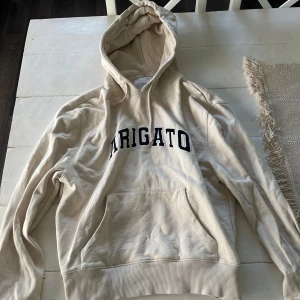 Beige hoodie från Axel Arigato - Säljer en beige hoodie från Axel Arigato med svart broderad text på bröstet. Tröjan har huva med snörning, stor magficka och ribbade muddar vid ärmslut och nederkant. Perfekt för chill dagar och enkel att matcha med jeans eller joggers. Osäker på storlek men tror det är en M.