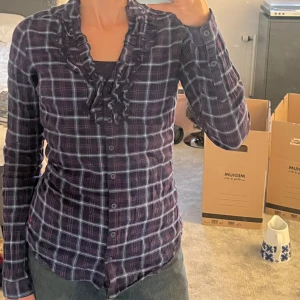 Rutig volang blus - Supersnygg rutig skjorta/blus från Fishbone i mörkblått, rött och vitt. Skjortan har volangdetaljer längs knäppningen och en broderad röd logga nertill. Långärmad och figurnära passform, med knappar längst bak så man kan knäppa åt den. 100% bomull.