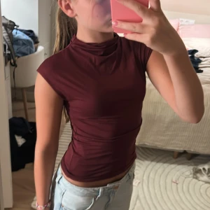 Vinröd polotopp från Gina Tricot - Säljer en vinröd polotopp från Gina Tricot i storlek XXS. Toppen är tight, har hög hals och är ärmlös med en slät, stretchig känsla. Perfekt att styla med jeans eller kjol för en snygg look.