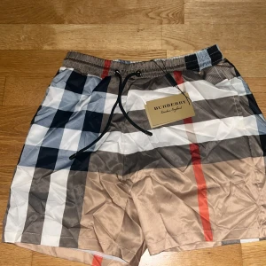 Rutig Burberry shorts med snörning - Snygga Burberry shorts med klassiskt rutigt mönster i beige, svart, vit och röd. De har elastisk midja med svart snörning och en bakficka. Materialet är lätt och känns som polyester, perfekt för sommaren. Ikonisk Burberry-tag medföljer s passform.