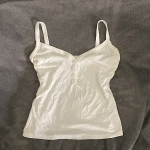 Vit ribbad linnetopp med knappar - Säljer en vit ribbad linnetopp med smala axelband och v-ringning framtill. Toppen har tre små knappar vid urringningen och är figurnära i modellen. Perfekt för varma dagar eller att styla i lager på lager.