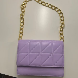 Lila quiltad axelväska med guldkedja - Säljer en lila quiltad axelväska med snygg guldfärgad kedja som axelrem. Väskan har ett stilrent rutmönster med trianglar och stängs med magnetlås. Perfekt storlek för det viktigaste och ger en poppig touch till din outfit.