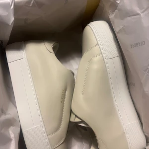 Beige sneakers från Axel Arigato - Stilrena sneakers från Axel Arigato i beige skinn med minimalistisk design och vit platt sula. Skorna har diskret logga i guld på sidan och saknar synlig snörning för en clean look. Perfekta för dig som gillar enkel och modern stil.