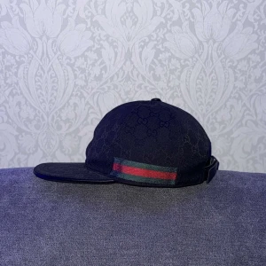 Svart Gucci keps med GG-mönster - Snygg svart keps från Gucci med klassiskt GG-monogram över hela kepsen och ikoniska gröna och röda ränder på sidan. Justerbar rem baktill för perfekt passform. Tillverkad i slitstark bomull och polyesterblandning.