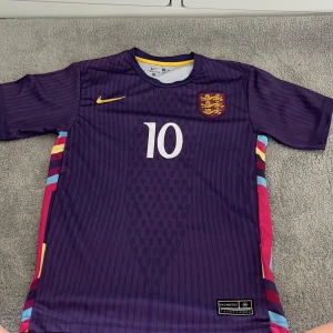 England Bellingham #10 fotbollströja Nike - Snygg England fotbollströja från Nike i mörklila med gula och röda detaljer. Nummer 10 och Bellingham på ryggen. Broderat England-emblem och Nike-logga. Tröjan har korta ärmar och är tillverkad i lätt, ventilerande material. Färgglada ränder längs sidorna för extra stil.