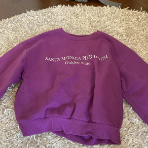 Lila sweatshirt från Gina Tricot - Cool lila sweatshirt från Gina Tricot med trycket 'Santa Monica Pier Hotel Golden State' på bröstet. Tröjan har rund hals, ribbade muddar och är långärmad. Perfekt för dig som gillar streetstyle och vill ha en färgstark look.