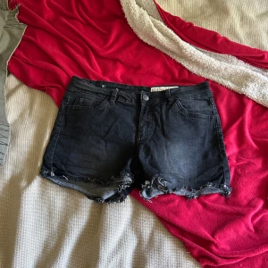 Svarta jeansshorts från Esmara - Svarta jeansshorts från Esmara i regular waist-modell. De har klassisk femficksdesign, råa och slitna kanter samt knapp och dragkedja framtill. Perfekta för dig som gillar en edgy och avslappnad stil.