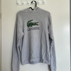 Grå hoodie från Lacoste med logga - Snygg grå hoodie från Lacoste med den klassiska gröna krokodilloggan och broderad text på bröstet. Tröjan har huva och långa ärmar, perfekt för chill dagar. Mjuk bomullskänsla och relaxed fit. 