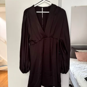 Svart kort klänning med v-ringning - Snygg svart klänning från gina med djup v-ringning och långa puffärmar. Tillverkad i ett glansigt satinliknande material som ger en lyxig känsla. Klänningen är kort och har en markerad midja för en smickrande siluett. Perfekt för dig som gillar stilrena och trendiga plagg.