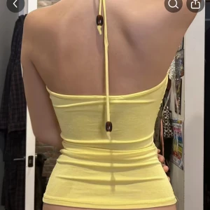 Gul halterneck topp med volang - Supersöt gul halterneck topp med volangdetaljer vid bysten och djup v-ringning. Toppen har smala knytband i nacken med träpärlor och är figurnära i modellen. Perfekt för varma dagar och ger en fräsch vibe.