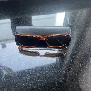 Bruna rektangulära Fedon solglasögon  - Snygga solglasögon med rektangulär form och brunmelerad båge. Glasen är mörka och bågen har en klassisk, stilren look. Kommer med svart fodral med gulddetalj. Perfekt för dig som vill ha en cool och tidlös accessoar. 