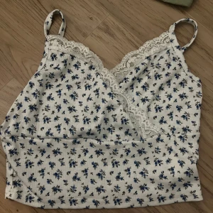 Blommig vit och blå linnetopp med spets - Supersöt linnetopp med små blå blommor på vit botten och fina spetsdetaljer längs v-ringningen. Tunt ribbat material och smala axelband ger en lätt och somrig känsla. Perfekt att matcha med jeans eller kjol för en fräsch look.