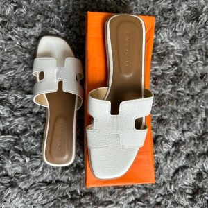 Vita sandaler med fyrkantig tå - Snygga vita sandaler från KIMY JIMMY med fyrkantig tå och platt sula. Ovandelen är i strukturerat skinn med ett coolt utskuret H-mönster. Perfekta för dig som gillar minimalistisk och trendig stil. Superlätta att bara glida i och dra iväg.
