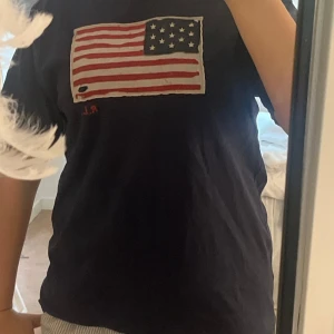 Svart t-shirt med amerikansk flagga RL - Mörk blå raulph lauren t shirt i storlek XL(18-20år) med en flagga vid bröstet! Hålet i flaggan är design och såg ut så när vi köpt den!💓