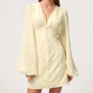 Gulblommig kortklänning från Adoore - Supersöt kortklänning från Adoore i ljusgul nyans med små gula blommor. Klänningen har v-ringning, långa puffärmar och knappar framtill. Säljer då jag köpte fel storlek