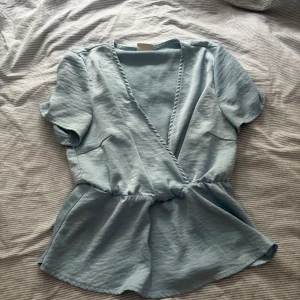 Ljusblå omlottopp med kort ärm - Supersöt ljusblå omlottopp med korta ärmar och markerad midja. Toppen har en v-ringad omlottfront och ett flowigt fall nedtill. Perfekt för dig som gillar en avslappnad men ändå trendig look.