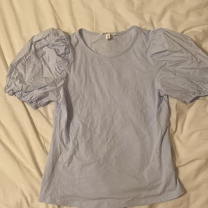 Ljusblå topp med puffärmar från H&M - Supersöt ljusblå topp från H&M med puffiga kortärmar och rund halsringning. Tillverkad i mjuk bomull, perfekt för dig som gillar en feminin och trendig stil. Enkel att matcha med både jeans och kjol.