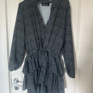 Svart prickig kortklänning Chicaca - Supersöt svart kortklänning från Chicaca med vita prickar, v-ringning och knappar framtill. Klänningen har långa ärmar, volangdetaljer och knyt i midjan för en snygg siluett. Perfekt för dig som gillar lekfulla mönster och trendiga detaljer.