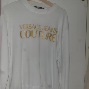 Vit sweatshirt Versace Jeans Couture - Säljer en clean vit sweatshirt från Versace Jeans Couture med guldigt tryck på bröstet. Tröjan har rund hals, långa ärmar och ribbade muddar. Perfekt för dig som vill ha en snygg och lyxig streetwear-look med statement. S passar även M.