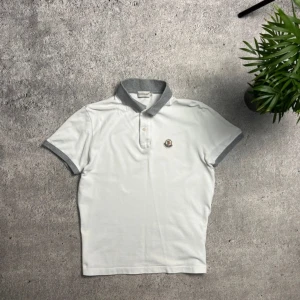 Vit Moncler pikétröja med grå krage - Vit pikétröja från Moncler med grå krage och gråa ärmslut. Klassisk Moncler-logga på bröstet och två knappar vid halsen. Tillverkad i bomull för en skön och luftig känsla. Perfekt för dig som gillar stilren och sportig look. Den har två små hål men som knappt syns. Passar M/S