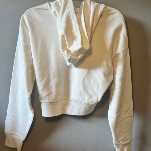Vit croppad hoodie från Lindex - Säljer en vit croppad hoodie från Lindex med huva och half zip-dragkedja i ryggen. Tröjan har långa ärmar och breda muddar vid ärmslut och nederkant. Perfekt för dig som gillar enkel och clean stil. Materialet är mjuk bomull som känns skönt mot huden.