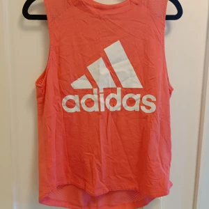 Korallfärgad Adidas linnetopp - Sportig korallfärgad linnetopp från Adidas med stort vitt logotryck på framsidan. Toppen är ärmlös och har mesh-paneler baktill för extra ventilation. Perfekt för dig som gillar att träna och vill ha en snygg och bekväm look.