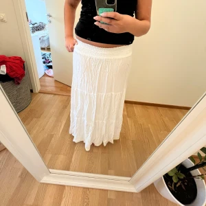 Vit långkjol med smockad midja - Säljer en vit långkjol med smockad midja och lätt rynkad struktur. Kjolen har en flowy och luftig känsla, perfekt för varma dagar. Materialet känns som bomull och kjolen har en enkel, stilren look utan mönster eller extra detaljer.