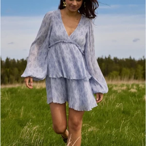 Ljusblå omlottklänning med volanger - Superfin ljusblå kortklänning med v-ringning och omlottliv. Klänningen har långa, luftiga ärmar och dubbla volanger i kjolen som ger en flowy känsla. Tunt, skönt material med diskret blommigt mönster. Perfekt för vår och sommar, matcha med sneakers eller sandaler.