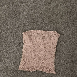 Ljusrosa smockad bandeau tubtopp - Supersöt ljusrosa bandeau tubtopp med smockad struktur och volangkant upptill och nedtill. Perfekt för varma dagar och enkel att styla med jeans eller kjol. Toppens stretchiga material gör den bekväm och följsam.