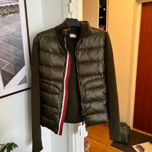 Olivgrön dunväst från Moncler - Snygg olivgrön dunväst från Moncler med stickade ärmar och quiltad front. Västen har hög krage, dragkedja med rödvit detalj och ribbade muddar. Materialmix av dun och stickad ull ger både värme och stil. Perfekt för lager-på-lager och streetwear. Det är en XL men sitter som en L, jag har vanligtvis M och såhär sitter den på mig^^