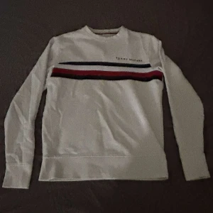 Vit långärmad tröja Tommy Hilfiger - Snygg vit långärmad tröja från Tommy Hilfiger med klassiska ränder i blått, rött och svart över bröstet. Tröjan har rund hals och är tillverkad i mjuk bomull, perfekt för chill dagar. Diskret logga på framsidan ger en clean look.