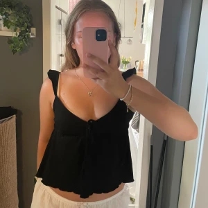 Svart linnetopp med volang från Nelly - Supersöt svart linnetopp från Nelly med volangdetaljer på axelbanden och lätt v-ringning. Toppen har knyt framtill och är croppad med flowig passform🩷