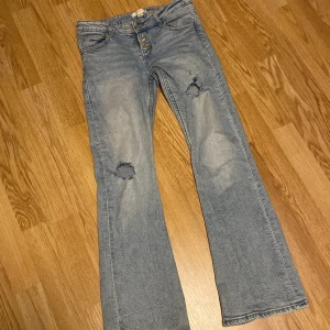 Ljusblå bootcut jeans med slitningar - Säljer ett par ljusblå jeans med bootcut-modell och slitna detaljer på båda benen. Jeansen har klassisk femficksdesign, knappgylf och är tillverkade i mjukt denimtyg. Perfekta för en avslappnad och trendig look.