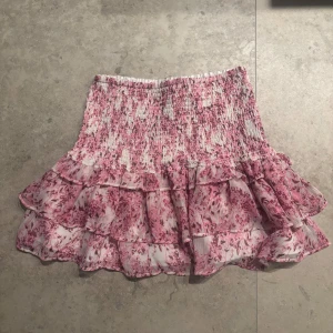 Rosa volangkjol med blommigt mönster från Gina - Supersöt kort kjol i rosa och vitt med blommigt mönster och flera volanger. 