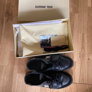 Axel arigato clean 90 svarta - Säljer ett par svarta sneakers från Axel Arigato med snygg broderad fågel på sidan. Skorna har snörning, rund tå och platt sula. Tillverkade i skinn med detaljer i mocka framtill. Kommer med originalkartong och extra skosnören.