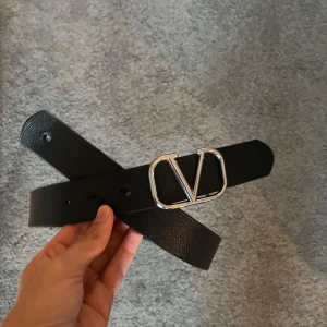 Svart Valentino bälte med silverdetalj - Stilrent svart bälte från Valentino med bred rem i skinn och en stor, ikonisk silverspänne med V-logga. Perfekt accessoar för att lyfta vilken outfit som helst med en lyxig touch.