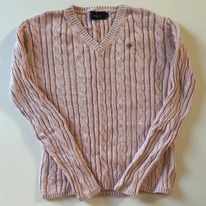 Ljusrosa kabelstickad tröja Morris - Säljer en ljusrosa kabelstickad tröja från Morris Lady i storlek XS. Tröjan har v-ringad hals, långärmade och ett litet broderat emblem på bröstet. Mjuk och skön kvalitet, perfekt för dig som gillar klassisk stil med en modern touch.