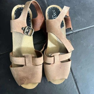 Bruna träskosandaler från Sweeks - Snygga sandaler från Sweeks i ljusbrunt skinn med träklack och nitar längs sidorna. Remmarna har en cool spänndetalj i metall och öppen tå. Perfekta för sommarens alla outfits och ger en retrovibe med sin träbas och klassiska design.