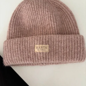 Rosa ribbad mössa från Marta du Château - Mysig ribbstickad mössa i ljusrosa från Marta du Château. Klassisk modell med uppvikt kant och diskret logga framtill. Tillverkad i en mjuk mix av polyester, ull och akryl som håller dig varm under kyliga dagar.