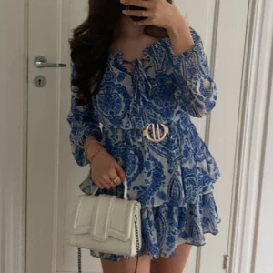 Blåvit kortklänning med volanger - Säljer en superfin blåvit kortklänning med långärmade ärmar och snygg snörning vid halsen. Klänningen har ett vackert blommigt mönster, volanger och ett dekorativt bälte i midjan. 💙