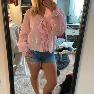 Rosa volangblus från Nelly - Supersöt ljusrosa blus från Nelly med volangdetaljer framtill och på ärmarna. Långärmad modell med knytband vid handlederna och lätt skrynkligt tyg. Perfekt för dig som gillar romantisk och trendig stil.